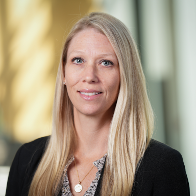 Rebecca McNally Keehn poses for her headshot. She is a fair skinned woman with blonde hair, wearing a black blazer over a multicolored blouse.