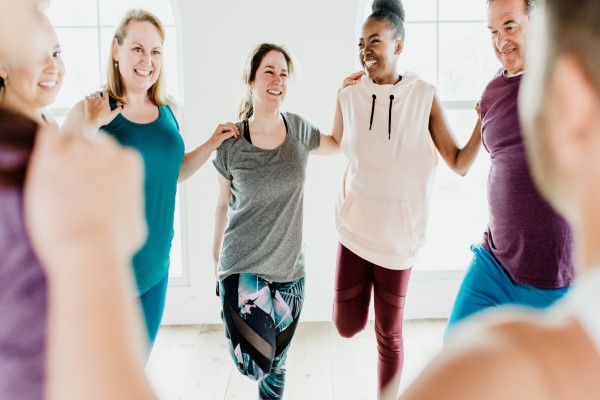 A diverse group of women dressed in athleisure clothing stand arm in arm as if they are in the middle of group stretching exercises.