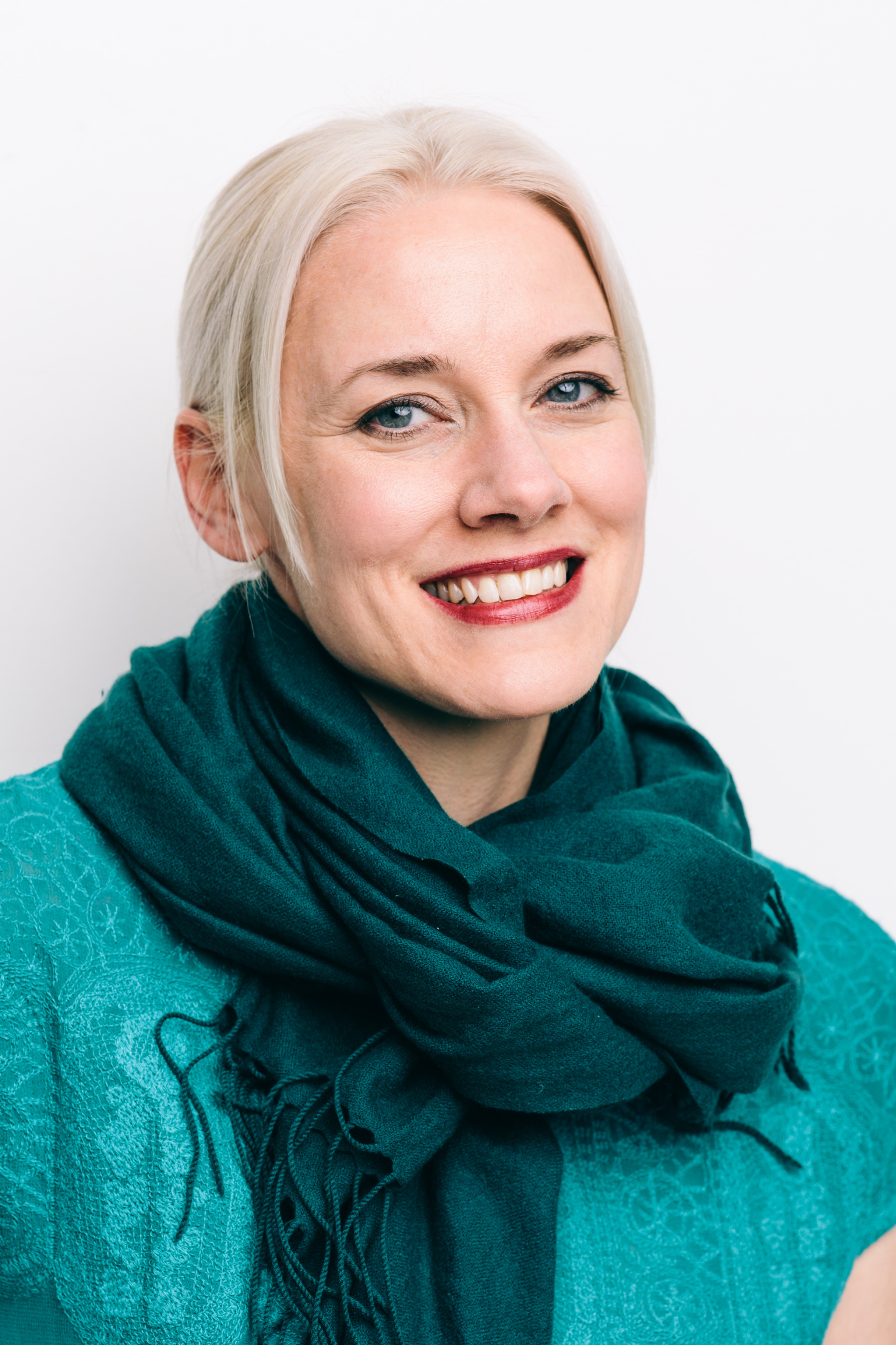 Dr. Heather Eicher-Miller smiles in a headshot. She is a white woman with white blonde hair swept back into an updo, wearing a bright teal shirt and a turquoise scarf around her neck.