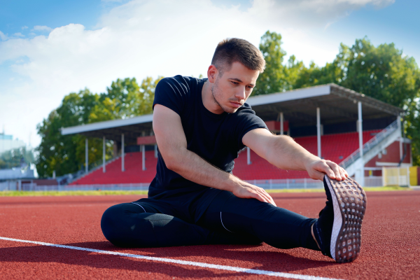 A man does a sitting hamstring stretch on a running track in front of empty stands.