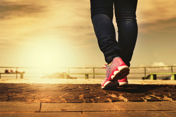 a pair of legs wearing what appears to be jeans and bright pink tennis shoes walks from the foreground of the image towards the background. The background appears to be a beachy landscape with railing and a setting sun that has drenched everything in golden light.